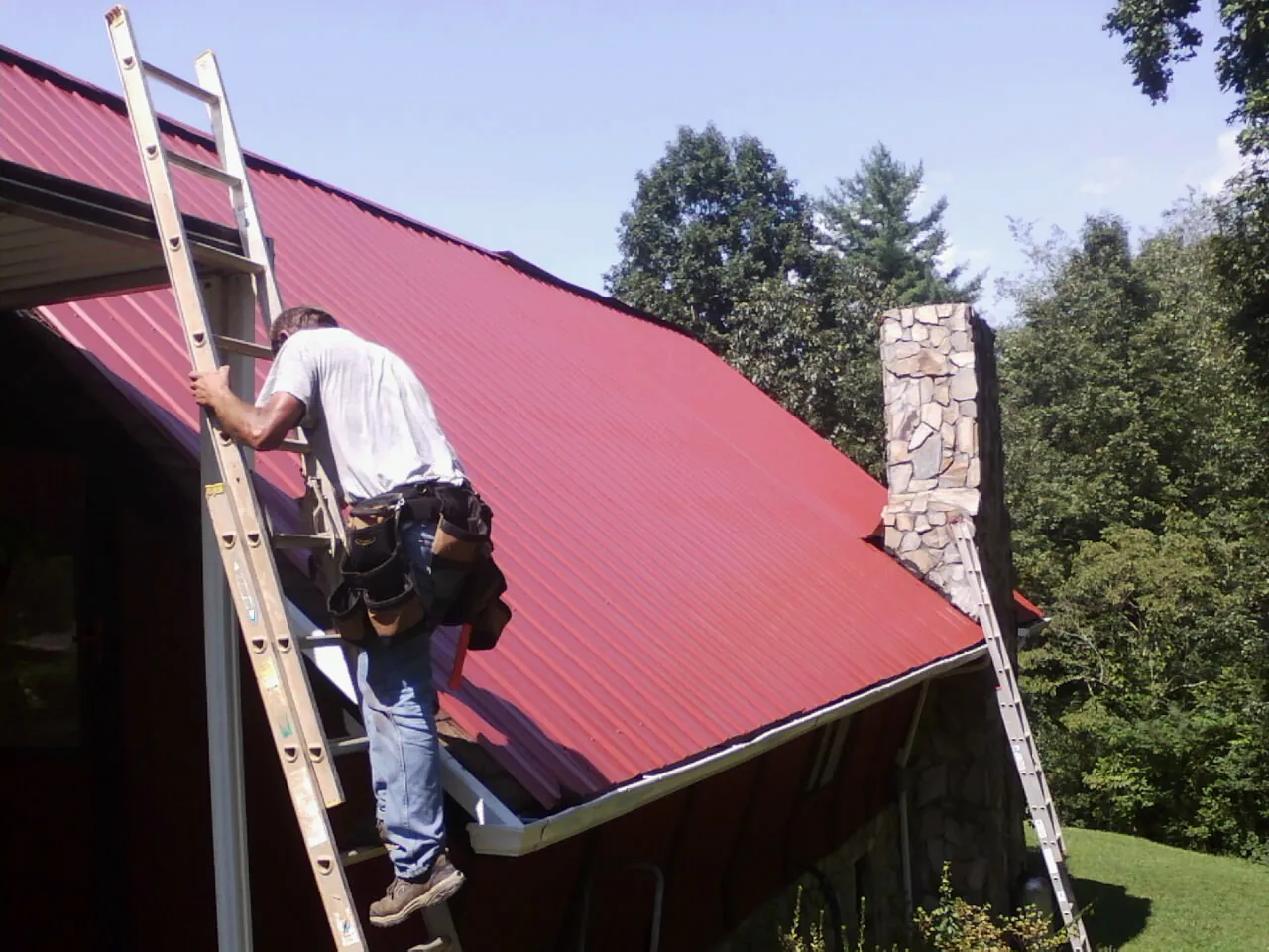 Roofer on a ladder beside a red metal roof and stone chimney — Soffit & Fascia Repair by Roofing Brothers in Essig
